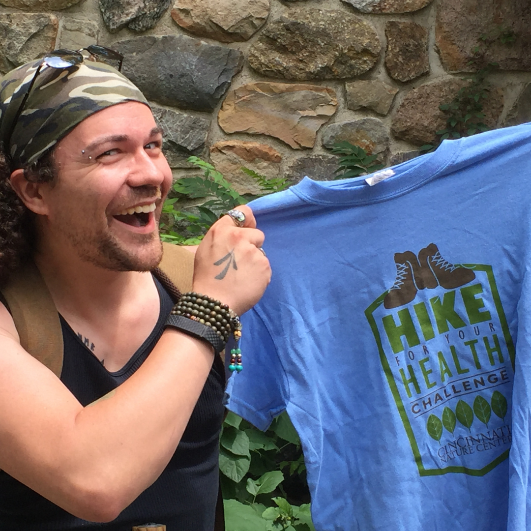 A young white man smiles at the camera as he holds up a Hike for Your Health t-shirt